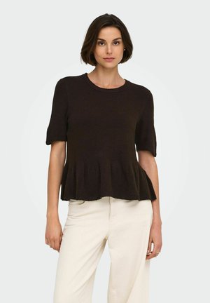 Femme aux cheveux courts et foncés portant un haut péplum noir à manches courtes et un pantalon beige clair, debout devant un fond uni.