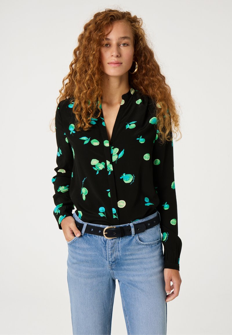 Schwarze Bluse mit grünen Blatt- und Fruchtmustern, mit Kragen, Knopfleiste und langen Ärmeln. Kombiniert mit hellblauen Jeans und einem Gürtel.