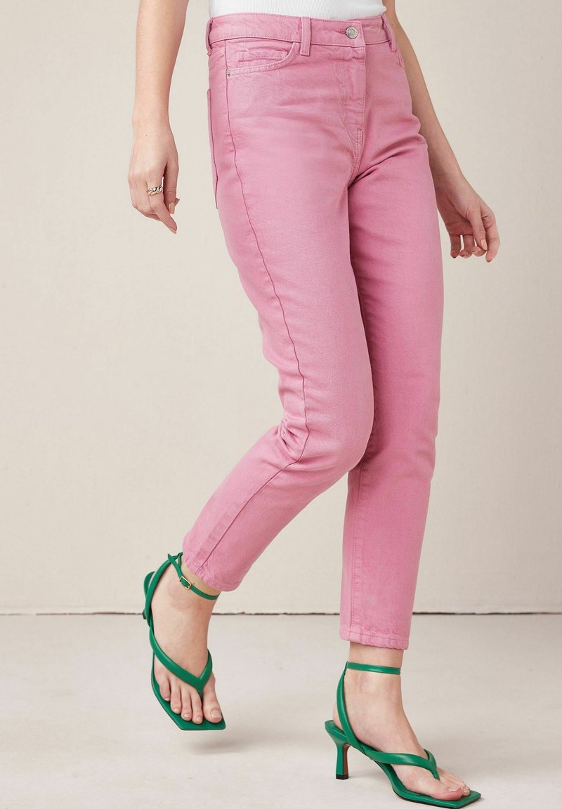 Jean slim, rose clair, avec une légère extensibilité, design à cinq poches et une texture subtile, associé à des sandales à talons verts à lanières.