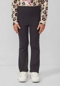 Enfant portant un pantalon noir côtelé, un haut à manches longues imprimé léopard et des baskets blanches, se tenant contre un fond gris uni.