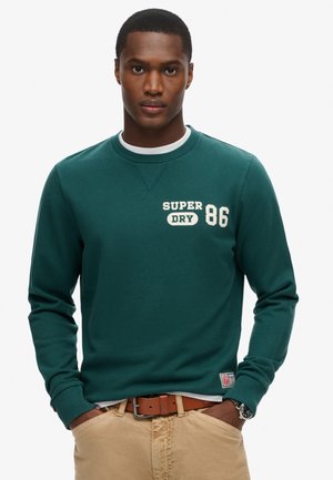 Green cotton sweatshirt with ribbed cuffs and crew neckline. Features cream-colored printed logo "SUPER DRY 86" on the front.