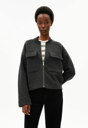 Frau mit kurzen lockigen Haaren, die eine dunkelgraue Jacke mit Reißverschluss und großen Taschen sowie schwarze Hosen trägt, steht vor einem weißen Hintergrund.