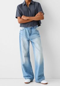 Oříznutá tmavě šedá košile na knoflíky, široké světle modré džínové kalhoty s vybledlou texturou a bílé tenisky s hnědými podrážkami.