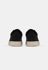 Paire de baskets basses noires avec semelles épaisses beige, vues de dos sur un fond clair uni.