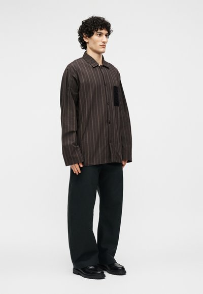 Άνδρας με σγουρά μαλλιά φοράει υπερμεγέθη καφέ πουκάμισο με ρίγες, μαύρο παντελόνι φαρδύ και μαύρα παπούτσια, στέκεται μπροστά σε απλό φόντο.