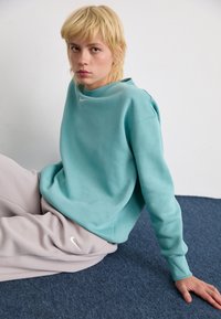 Ung person med kort blondt hår sidder på et blåt tæppe iført en teal sweatshirt og lysegrå Nike sweatpants, og ser ind i kameraet.