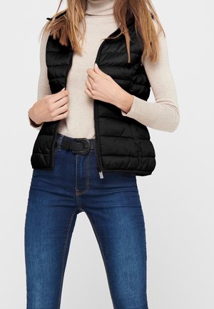 Vrouw draagt een beige coltrui, een zwarte doorgestikte mouwloze vest en blauwe spijkerbroek, met handen die de rits van het vest aanpassen.