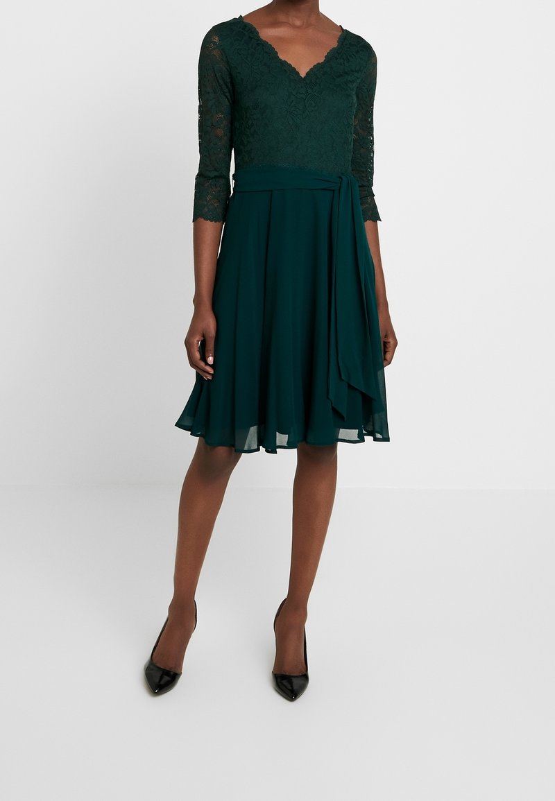 Mörkgrön klänning med spetsliv, V-ringning, 3/4 spetsärmar och en flödande, knälång kjol med knytskedja i midjan. Svarta klackar.