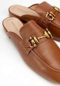 Scarpe slip-on in pelle marrone con una texture liscia. Presentano un'accento di fibbia dorata e dettagli di cucitura lungo i lati.