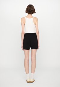 Shorts negros con una textura suave, de cintura alta, combinados con una camiseta blanca sin mangas. La modelo está de pie con la cabeza girada, mostrando un diseño simple.