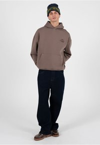 Mann trägt einen braunen Kapuzenpullover, dunkle lockere Jeans, braune Schuhe und eine schwarze Mütze mit gelbem Schriftzug, steht vor einem schlichten weißen Hintergrund.