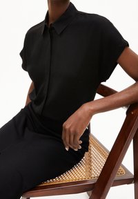 Camicetta nera a maniche corte con bottoni e texture morbida, indossata con pantaloni neri; soggetto seduto su una sedia di legno con seduta intrecciata.