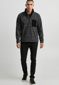 Grijze fleece pullover met een half-rits kraag, voorzien van een zwarte zak met rits op de borst. Draagt met zwarte broek en sneakers. Soepele textuur.