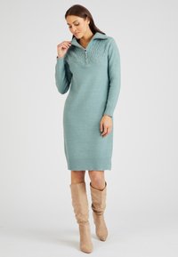 Abito lavorato a maglia di colore turchese chiaro con colletto a mezza zip e pannello superiore testurizzato. Presenta maniche lunghe e una silhouette aderente, abbinato a stivali beige.