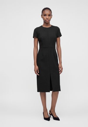 Frau steht in einem schwarzen knielangen Kleid mit kurzen Ärmeln und Vorder- Schlitz, trägt schwarze Slingback-High-Heels vor einfarbig grauem Hintergrund.