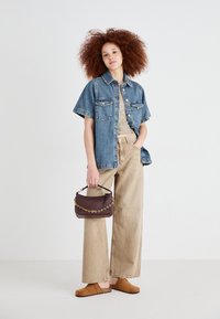 Jeans jakna s kratkimi rokavi preko bež kratkega topa, v kombinaciji z širokimi bež hlačami in rjavimi natikači, drži pleteno temno torbo.