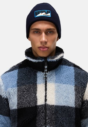 Fleece jacket with a black, blue, and white checkered pattern, featuring a high collar and zipper. The navy beanie has a logo patch.