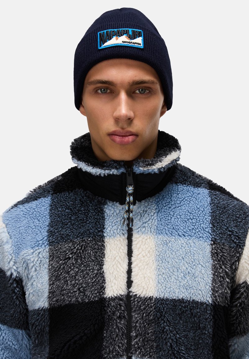 Fleece jacket with a black, blue, and white checkered pattern, featuring a high collar and zipper. The navy beanie has a logo patch.