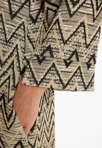 Strukturiertes Gewebe mit einem Zickzackmuster in Schwarz auf einem beigen Hintergrund; sichtbare Tasche am Ärmel des Kleidungsstücks.