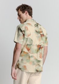 Korte mouwen shirt van een lichte stof met een beigekleurige basis en abstracte aquarelpatronen in groene en roestkleurige tinten. De achterkant heeft een kraag en knoop.