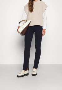 Beige Strickpullover mit kurzen Ärmeln, marineblaue enge Hose mit seitlichen Schlitzen und cremefarbene Loafers mit schwarzen Sohlen. Handtasche in Braun und Creme.