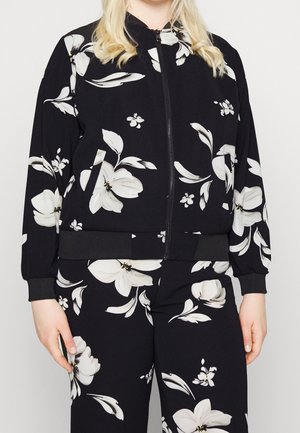 Frau trägt schwarze Jacke mit Reißverschluss und Hose mit großem weißem Blumenmuster, steht mit locker an den Seiten hängenden Armen.