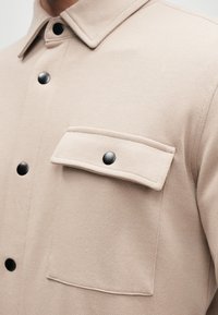 Giacca camicia beige chiaro in tessuto texture, con colletto strutturato, bottoni a pressione neri e una tasca a patta con chiusura a bottone.