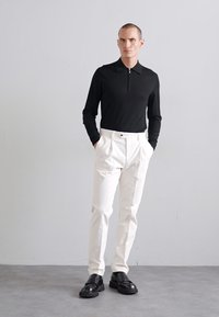 Camisa polo preta de mangas longas com fecho, combinada com calças brancas de cintura alta. A modelo usa sapatos pretos robustos, posicionando-se contra um fundo claro.