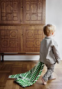 Barn i grå tøj, der går væk og trækker et hvidt stof med grønne prikker. Træskab med udskårne mønstre i baggrunden.