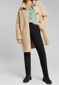 Beige fuskpälsjacka med krage, fem knappar, två fickor, bärs över en grön stickad topp och svarta byxor, kombinerat med svarta stövlar.