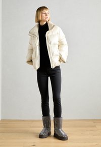 Pufferjakke i myk beige med ståkrage, kombinert med svarte tettsittende jeans og glitrende grå ankelstøvler. Glatt tekstur og skinnende detaljer.