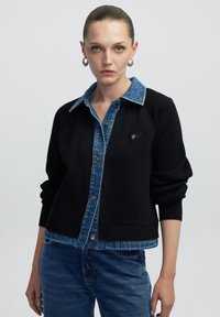 Zwarte cardigan met blauwe denim kraag, geribbelde textuur, knopen aan de voorkant en een klein driehoekig embleem op de borst. Casual pasvorm en ontwerp.