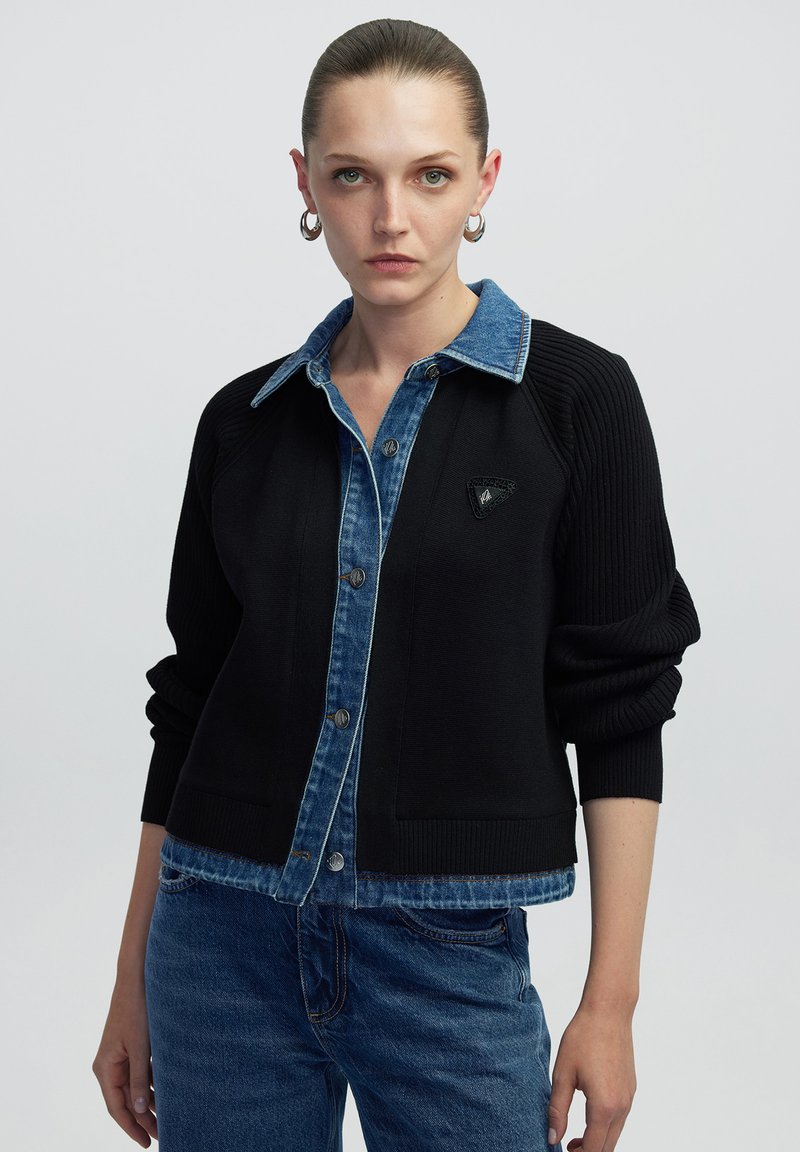 Zwarte cardigan met blauwe denim kraag, geribbelde textuur, knopen aan de voorkant en een klein driehoekig embleem op de borst. Casual pasvorm en ontwerp.