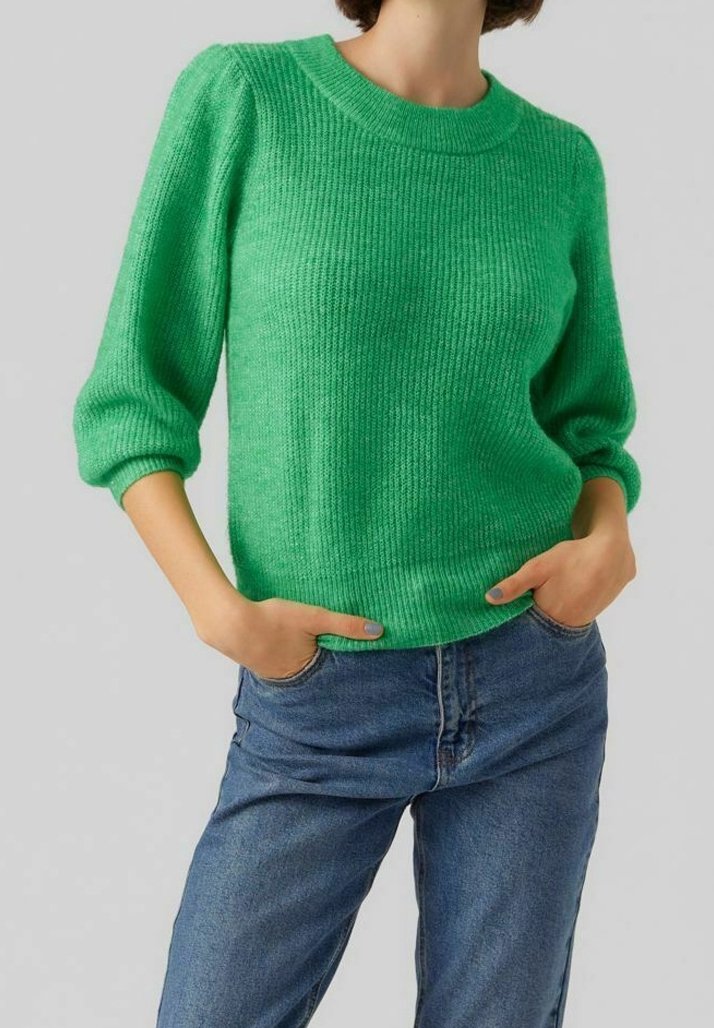 Personne portant un pull tricoté vert vif à manches trois-quarts et un jean bleu, les mains reposant dans les poches du jean.