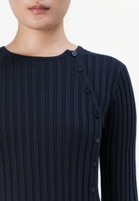 Navy gerippter Pullover mit Rundhalsausschnitt, strukturiertem Stoff und einer Knopfleiste entlang der linken Seite mit schwarzen Knöpfen.