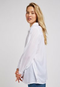 Camicia bianca a maniche lunghe con bottoni, realizzata in tessuto leggero. Presenta una vestibilità comoda, spacchi laterali e un design con colletto classico.