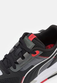 Baskets de sport noires et grises avec matériau en mesh, accents gris, détails rouges et surfaces texturées. Présente un logo sur la languette et les œillets.