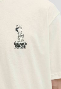 Lys beige bomulds t-shirt med et grafisk print af en tegneserie-mekaniker, der holder en skruenøgle. Teksten "CLASSIC GARAGE BRAKE BROS." står nederst.