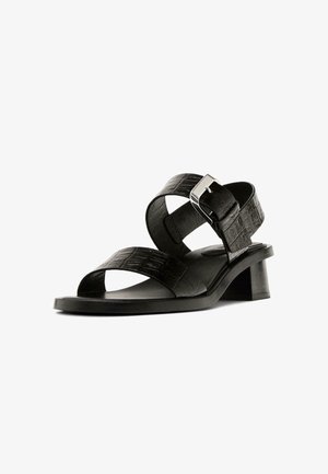 Schwarze Leder-Sandale mit zwei Kreuzriemen und einer silbernen Schnalle. Sie hat einen niedrigen Blockabsatz und eine glatte, flache Sohle mit strukturierter Oberfläche.