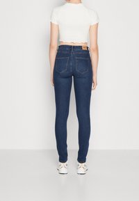 Modré džínové kalhoty s úzkým střihem a vysokým pasem, s viditelnými švy a dvěma zadními kapsami. Kombinovány s bílým crop topem a teniskami.