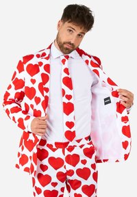Biely oblek s veľkými červenými srdcovými vzormi, zodpovedajúcou kravatou a nohavicami. Vnútorné podšívka je jednofarebná biela. Štandardný strih, s tradičnými klopami a gombíkmi.