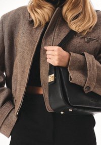 Nainen ruskeassa herringbone-takkissa, joka pitää mustaa nahkakäsilaukku kulkuset raskaudella, pukeutuneena mustiin yläosaan ja housuihin.