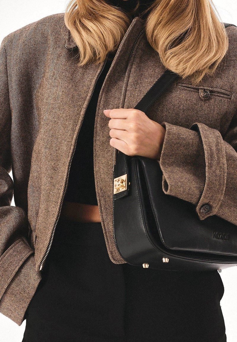 Nainen ruskeassa herringbone-takkissa, joka pitää mustaa nahkakäsilaukku kulkuset raskaudella, pukeutuneena mustiin yläosaan ja housuihin.