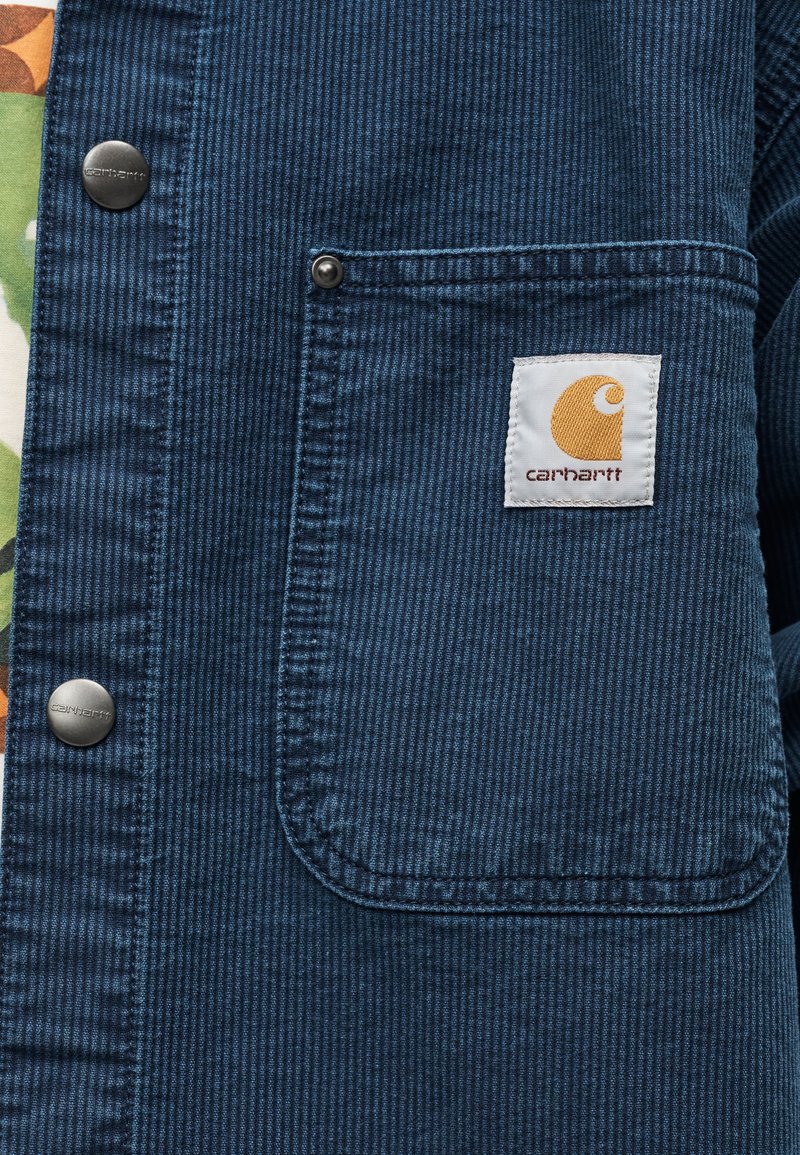 Blaue Cordjacke mit großer Fronttasche mit einem weißen Carhartt-Logo-Patch und metallischen Druckknöpfen an der Vorderseite.