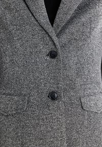 Grå strukturerad blazerväst med skräddarsydd design, två svarta knappar och sidofickor, med ett subtilt hundtandsmönster i svart och vitt.