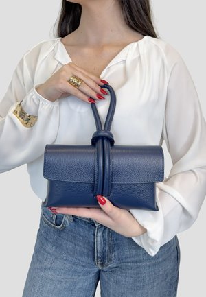 Donna con camicetta bianca e jeans che tiene una borsa in pelle blu scuro con manico ad anello, indossando un braccialetto d'oro e un anello, con unghie rosse.