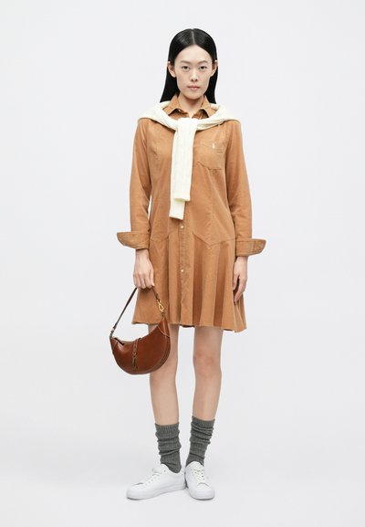 Καφέ φόρεμα από κορντούρο με εφαρμοστό μπούστο και φαρδιά φούστα. Λευκό πουλόβερ πλεκτό με σχέδιο κοτσίδες δέσμενο γύρω από τον λαιμό, γκρι κάλτσες και καφέ τσάντα χειρός.