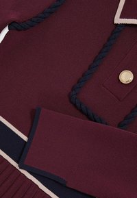 Kötött ruhadarab mély bordó színben, tengerészkék részletekkel. Zsinórozott díszítéssel és nagy arany gombbal rendelkezik. Puha textúrájú, bordázott elemekkel.