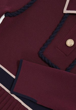 Stickad plagg i mörk burgundy med marinblå detaljer. Har flätade dekorationer och en stor guldknapp. Mjuk textur med ribbade inslag.