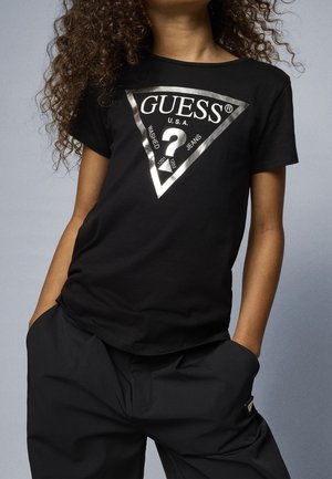 Schwarzes T-Shirt mit silbernem, dreieckigem GUESS-Logo mit dem Text "U.S.A. Washed Jeans", getragen von einer Person mit lockigem Haar und Händen in den Taschen schwarzer Hosen.